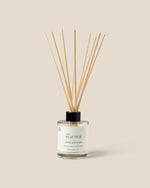 A reed diffuser bottle with a clear glass body, white label, black cap, and natural wood reeds, centered on a warm beige background. The label reads 'ACAI OUD' and 'AURA DIFFUSER'.