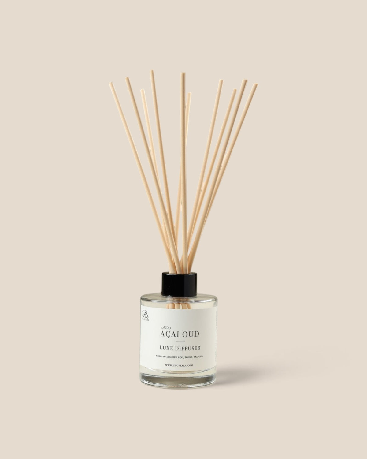 A reed diffuser bottle with a clear glass body, white label, black cap, and natural wood reeds, centered on a warm beige background. The label reads 'ACAI OUD' and 'AURA DIFFUSER'.