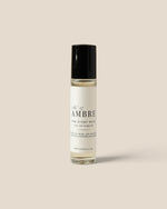 A roll-on perfume bottle with a black cap and a white label reading 'Ambre' , photographed against a solid beige background.