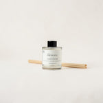 A clear glass reed diffuser bottle with a black cap and a white label reading 'FRESH FIG' and 'LUXE DIFFUSER', shown with several natural reed sticks on a clean off-white background.