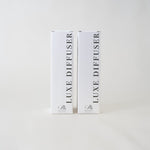 Two tall white rectangular product boxes standing upright on a light gray background. The boxes are labeled 'LUXE DIFFUSER' vertically in black text, with a small script logo near the bottom.
