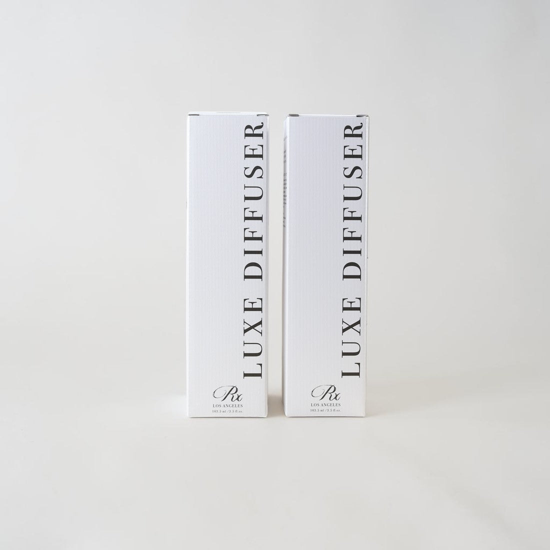 Two tall white rectangular product boxes standing upright on a light gray background. The boxes are labeled 'LUXE DIFFUSER' vertically in black text, with a small script logo near the bottom.