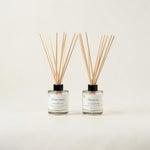 Two reed diffusers with clear glass bottles, white labels, and natural wooden reeds, set against a plain off-white background.