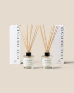 Two reed diffuser bottles with natural reed sticks and two tall white boxes labeled LUXE DIFFUSER, arranged on a warm beige background.