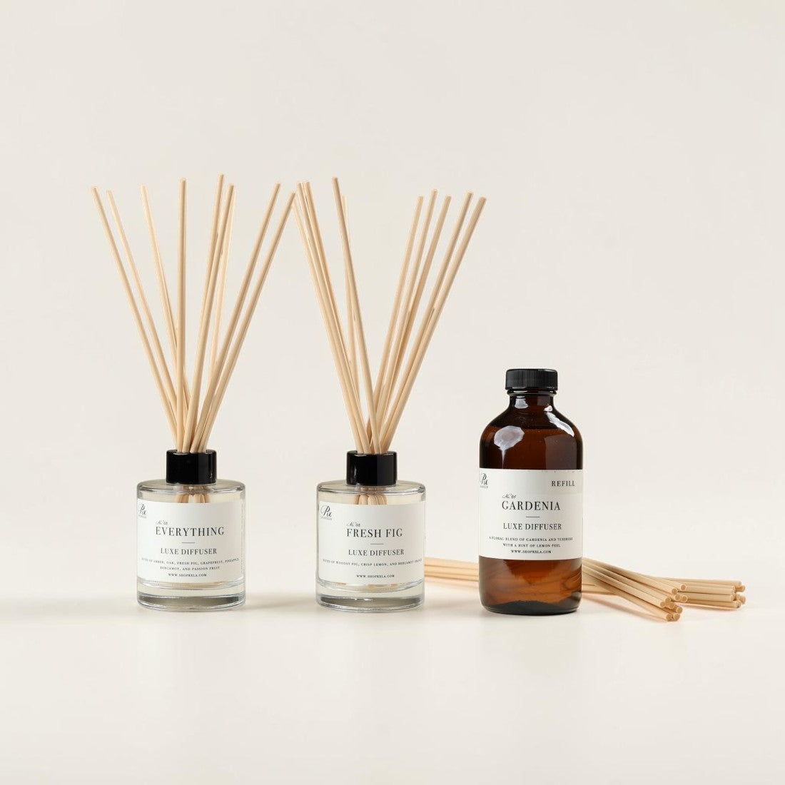 Two clear glass reed diffusers and one amber bottle with a white label on a clean white background. The diffusers have light natural reeds and black caps, and the bottle is labeled 'The Gardenia' reed diffuser.