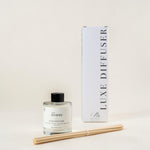A reed diffuser bottle with a black cap and clear glass, shown next to its tall white box labeled 'LUXE DIFFUSER' and two natural reed sticks on a warm off-white background.