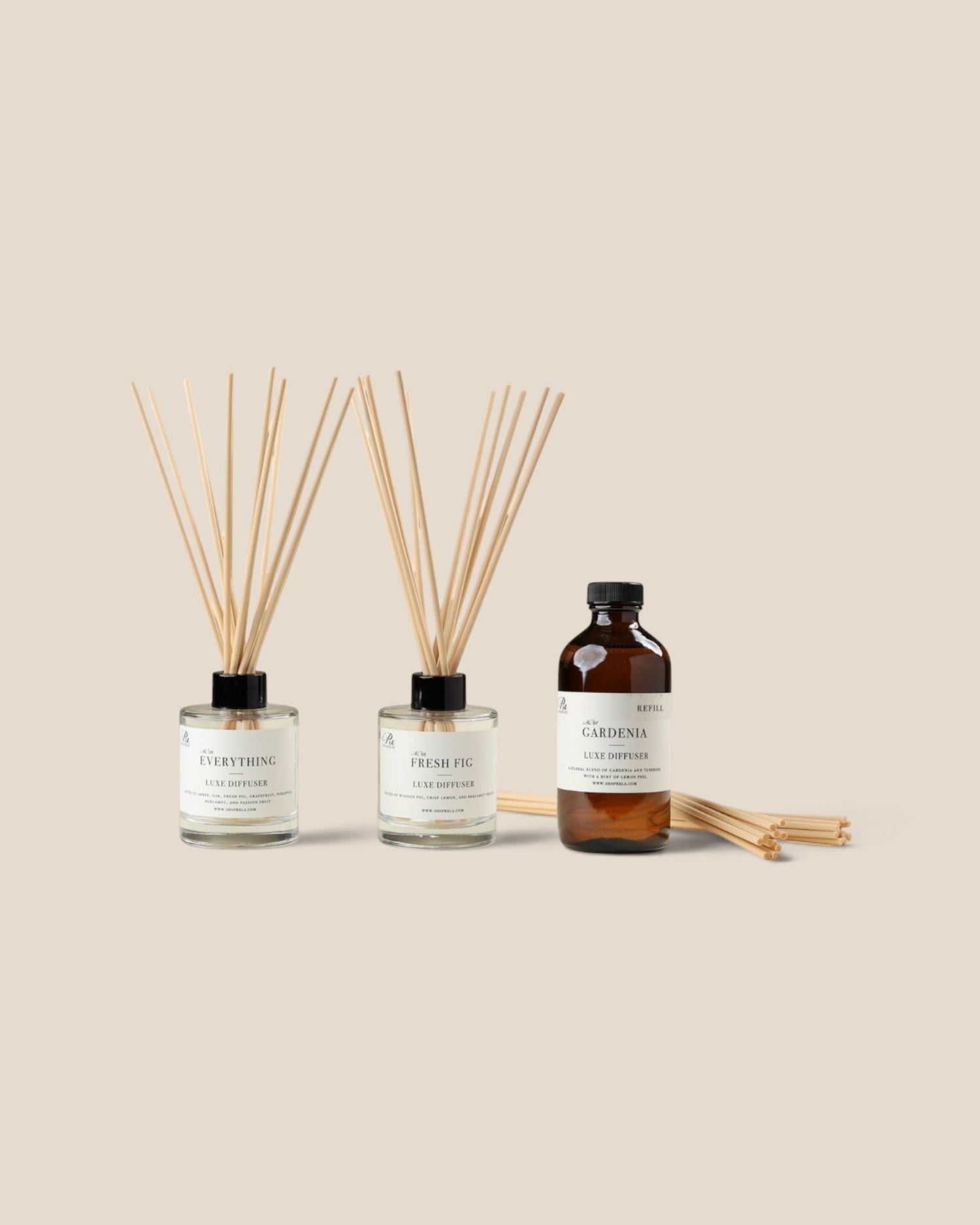 Three home fragrance products on a warm beige background: two clear glass reed diffusers with light wood reeds and one amber bottle with a white label and black cap.