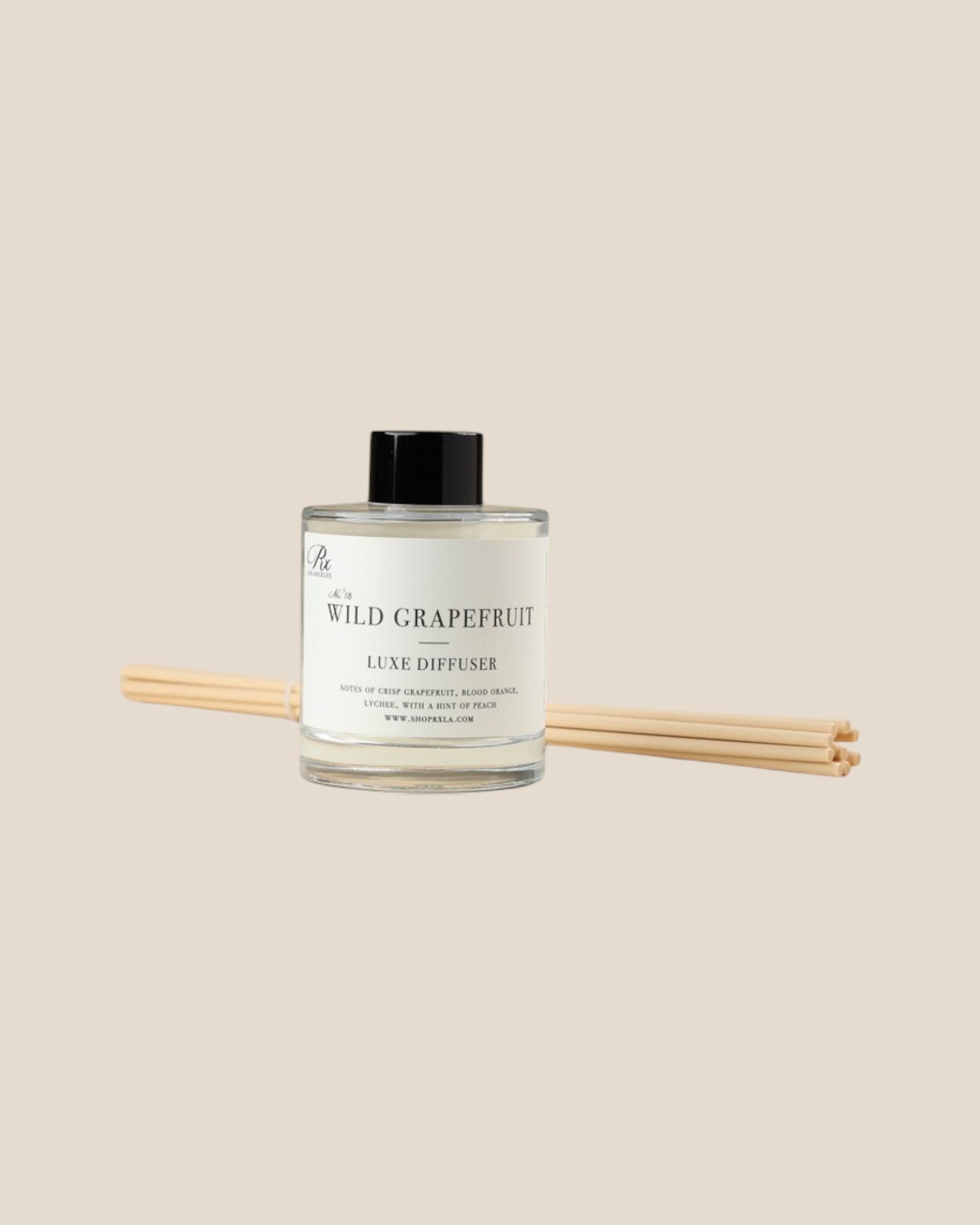 A reed diffuser bottle labeled 'Wild Grapefruit' with a black cap and reed sticks resting behind it, centered on a plain warm beige background.