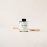 A clear glass reed diffuser bottle labeled 'An Earl Grey Luxe Diffuser' with a black cap, shown with two natural reeds on a plain light beige background.