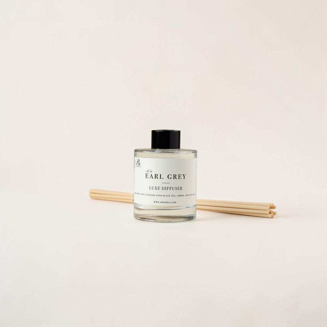 A clear glass reed diffuser bottle labeled 'An Earl Grey Luxe Diffuser' with a black cap, shown with two natural reeds on a plain light beige background.