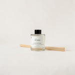 A clear glass reed diffuser bottle with a black cap and white label reading 'THE HOMME', shown with four light wood reeds on a plain white background.