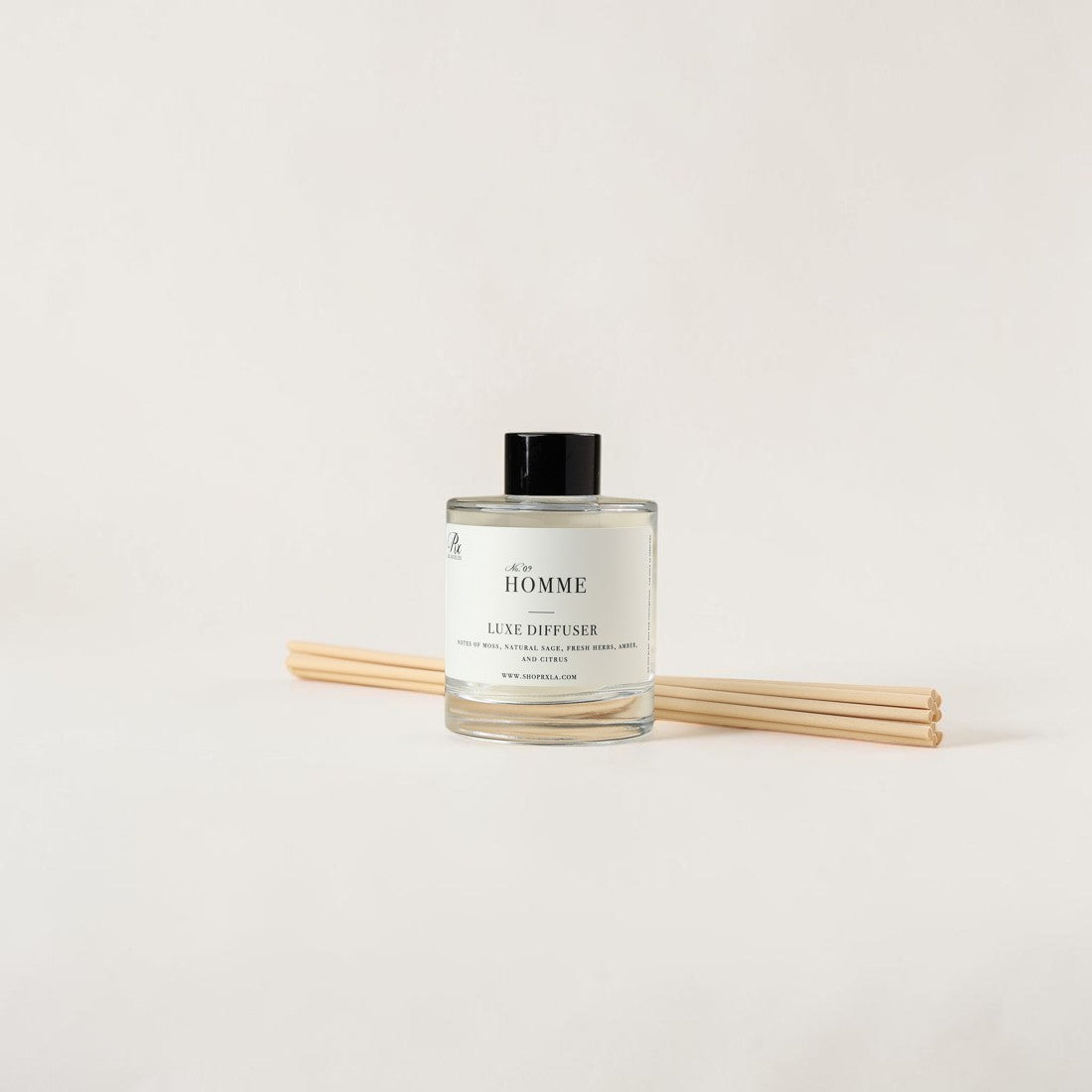 A clear glass reed diffuser bottle with a black cap and white label reading 'THE HOMME', shown with four light wood reeds on a plain white background.