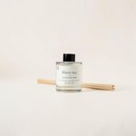 A clear glass reed diffuser bottle labeled 'THE WHITE TEA LUXE DIFFUSER' with a black cap, shown with two natural reeds behind it on a warm off-white background.