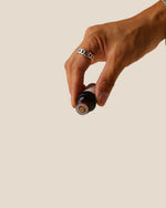 A close-up of a hand holding a small amber glass bottle with a black cap against a solid beige background.