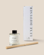A reed diffuser bottle with a black cap and white label next to its tall white box, with two natural reeds laid in front, on a plain beige background. The label reads 'LUXE DIFFUSER' 