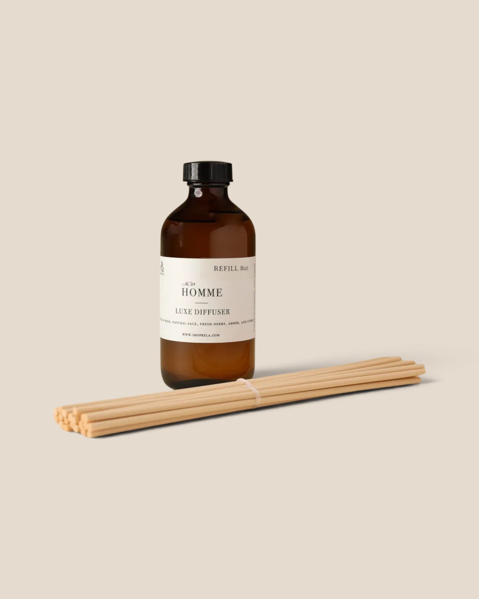 An amber glass bottle of Revill & Co. 'Le Homme' lit diffuser with a cream label and black cap, shown standing upright next to a bundle of natural wood diffuser sticks on a solid beige background.