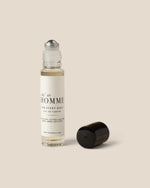 A clear glass rollerball perfume bottle with a white label reading 'HOMME' and a black cap placed beside it on a plain warm beige background.