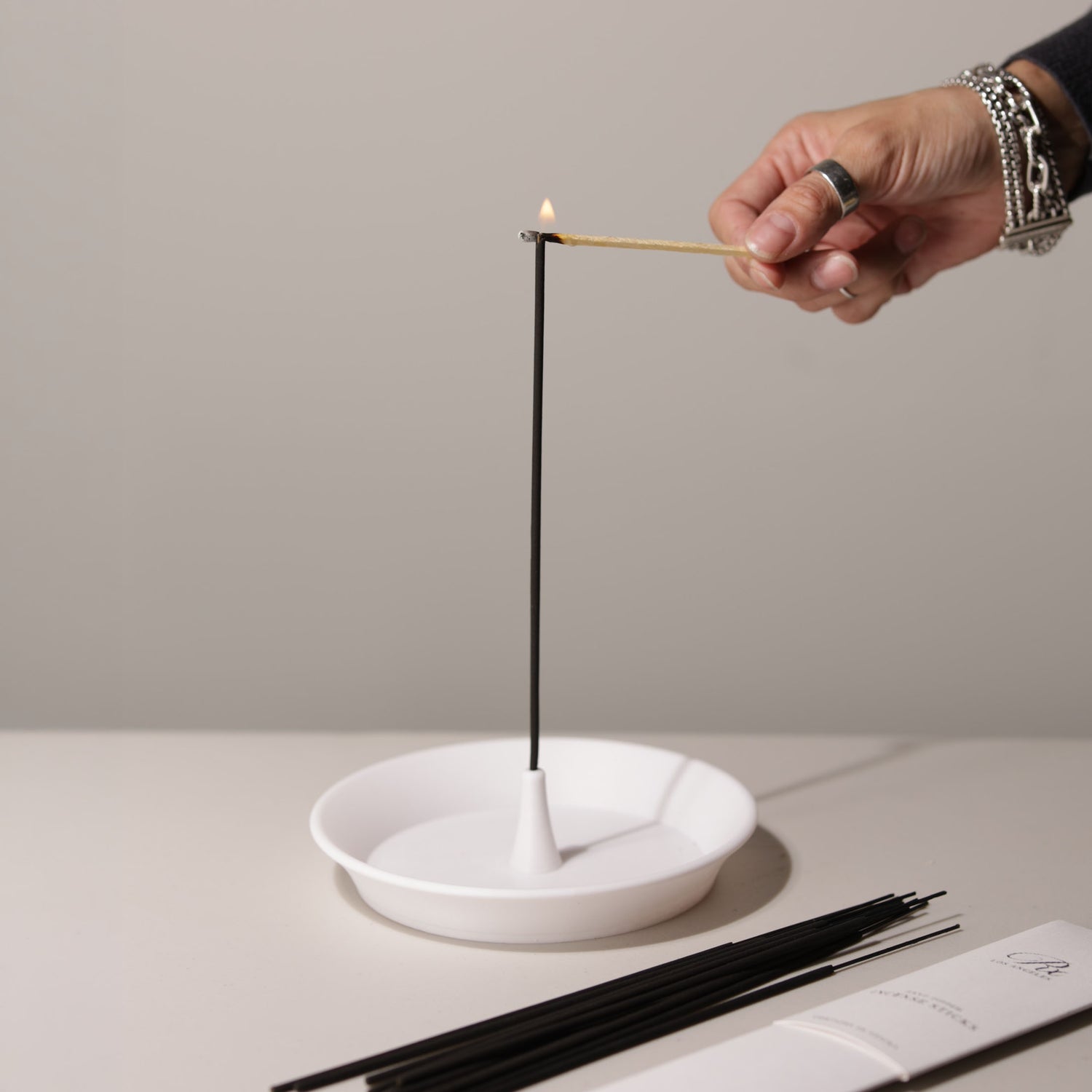 Person lighting an incense stick with a match, using a white ceramic holder on a neutral background.