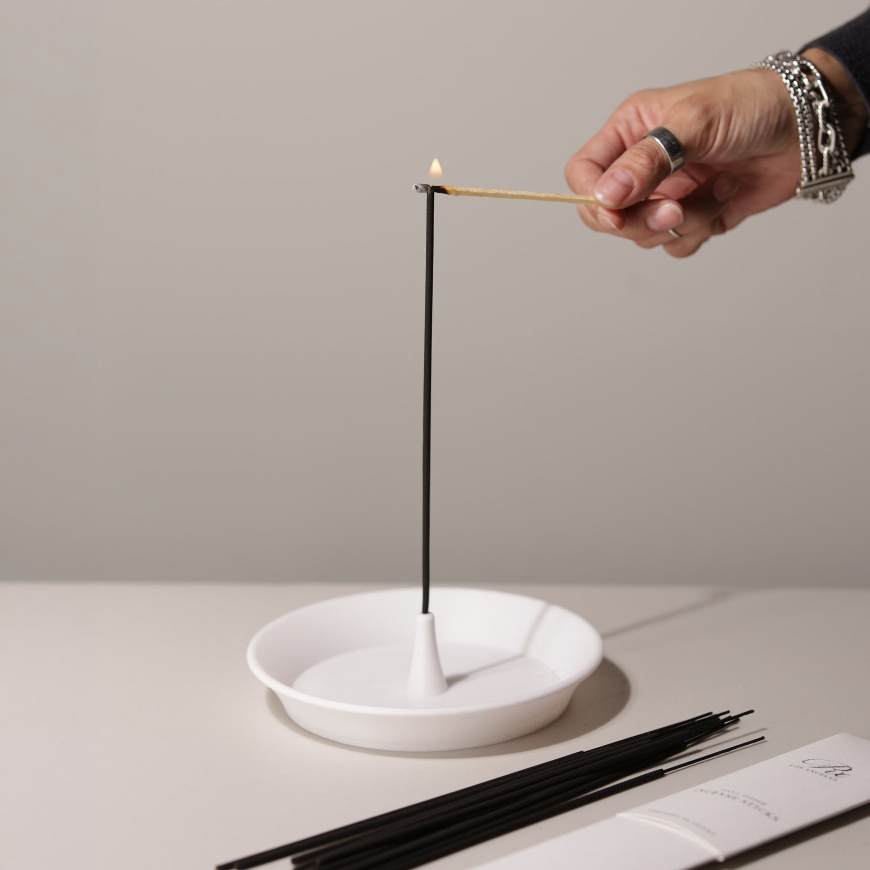 Person lighting an incense stick with a match, using a white ceramic holder on a neutral background.
