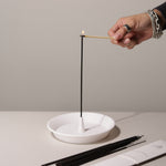 Person lighting an incense stick with a match, using a white ceramic holder on a neutral background.