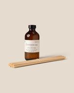 An amber glass bottle of Luxe Diffuser refill with a black cap and cream label, shown with a bundle of natural wood diffuser sticks on a solid beige background.