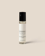 A clear glass rollerball perfume bottle with a white label and black cap, standing upright against a solid light beige background. The label reads 'LAVENDER CO FOR EVERY BODY EAU DE PARFUM'.