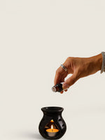 A hand holding a small essential oil bottle above a black ceramic candle warmer with a lit tealight candle, set against a plain light beige background.