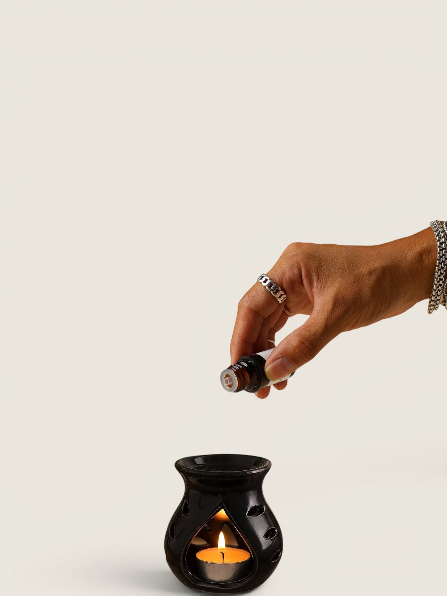 A hand holding a small essential oil bottle above a black ceramic candle warmer with a lit tealight candle, set against a plain light beige background.