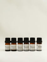 Six small amber glass fragrance oil bottles with black caps and white labels, lined up against a plain off-white background. The labels show different scent names 