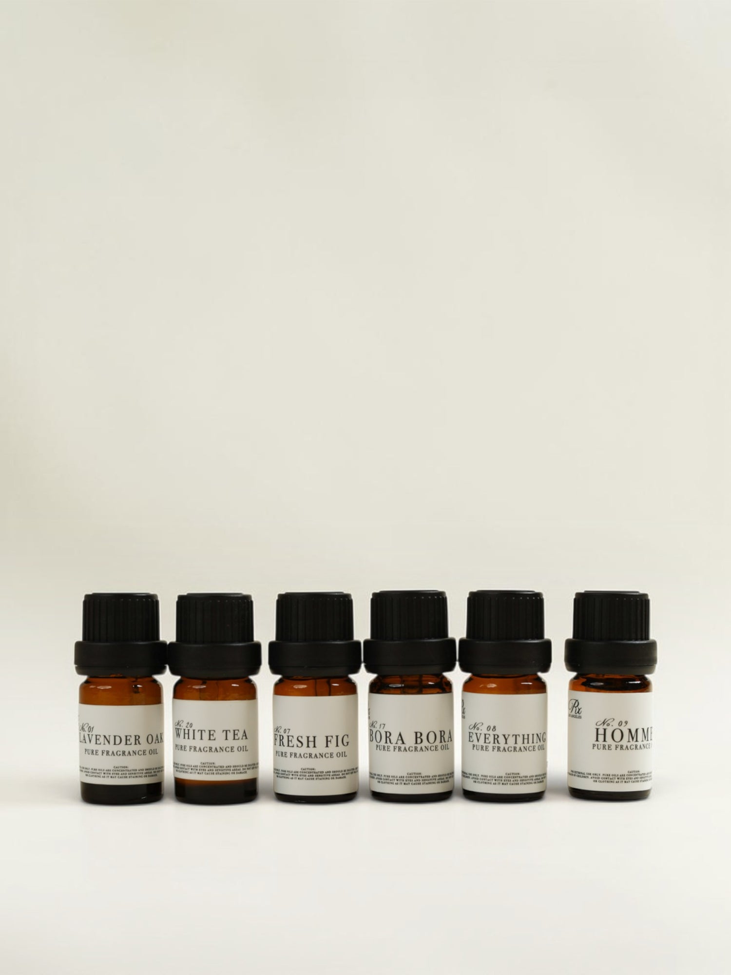 Six small amber glass fragrance oil bottles with black caps and white labels, lined up against a plain off-white background. The labels show different scent names 