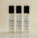 Three small clear perfume roller bottles with black caps and white labels, standing upright on a light beige background. The labels read No.