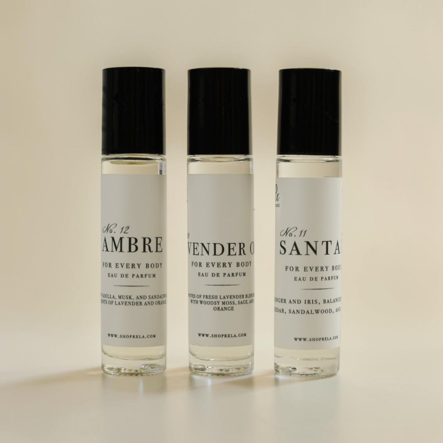 Three small clear perfume roller bottles with black caps and white labels, standing upright on a light beige background. The labels read No.