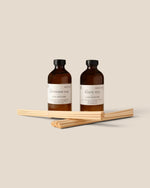 Two amber glass diffuser bottles with cream labels, one labeled Lavender Oak and the other White Tea, displayed with fiber diffuser sticks on a solid beige background.