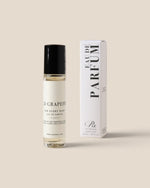 A clear glass roll-on perfume bottle with a black cap stands next to its white box packaging on a solid beige background. the box reads 'EAU DE PARFUM'.
