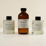 Three diffuser bottles arranged on a light beige background: two clear glass bottles with black caps and white labels reading 'White Tea Luxe Diffuser,' and one amber glass bottle with a black cap and a smaller white label.