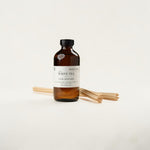 An amber glass bottle of Refill Co. White Tea Luxe Diffuser with a black cap and a white label, standing upright with several natural wood diffuser sticks behind it on a clean off-white background.
