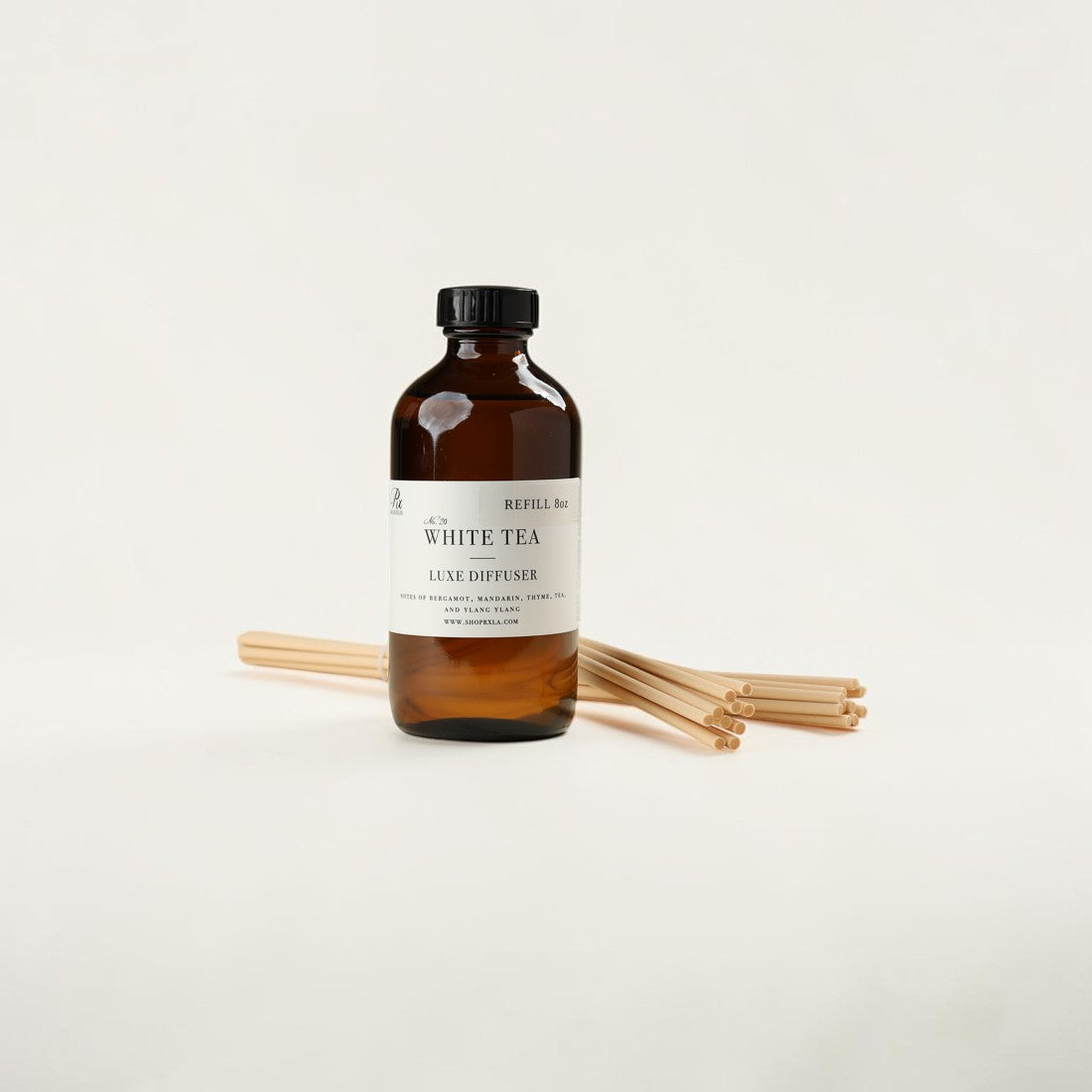 An amber glass bottle of Refill Co. White Tea Luxe Diffuser with a black cap and a white label, standing upright with several natural wood diffuser sticks behind it on a clean off-white background.