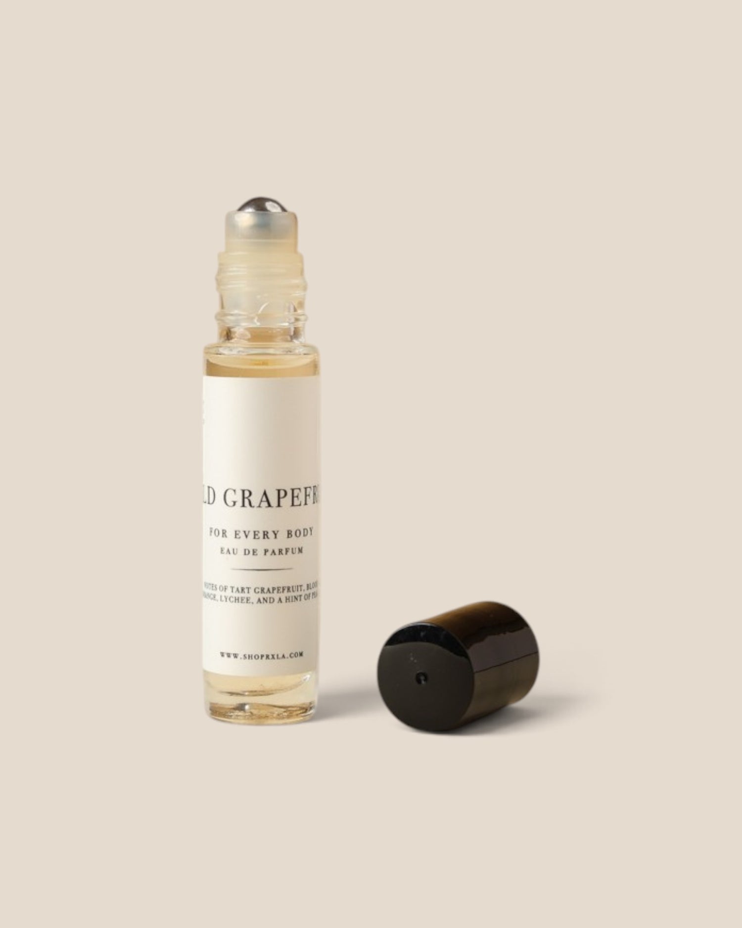 A clear glass rollerball perfume bottle with a white label reading 'wild grapefruit FOR EVERY BODY' stands next to its detached black cap on a warm beige background.